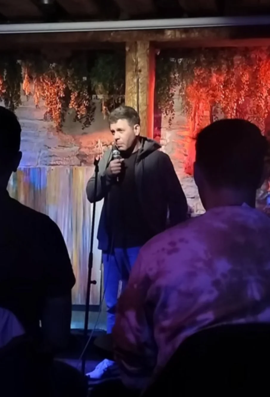 Artie Kay holding a microphone on stage at Winkel, with audience silhouettes in the foreground